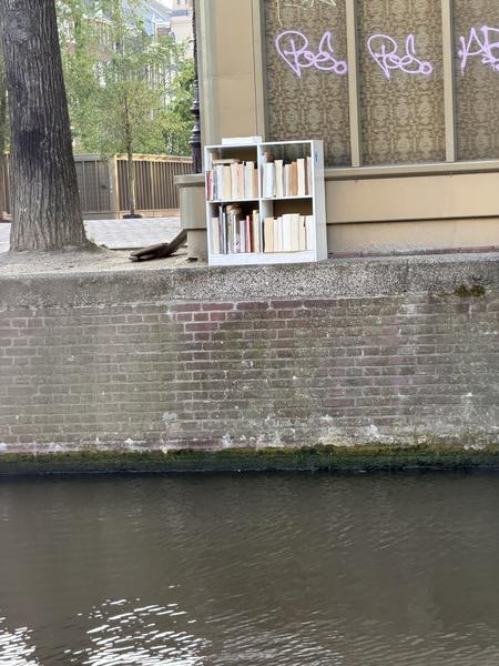 A small white bookshelf filled with books stands beside a canal in Centrum, Amsterdam, against a backdrop of a brick wall and a building with patterned windows
